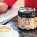 close up view of a person's hand writing on a piece of paper beside a cup of coffee and hemp extract on top of a table