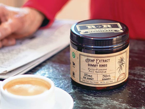 close up view of a person's hand writing on a piece of paper beside a cup of coffee and hemp extract on top of a table