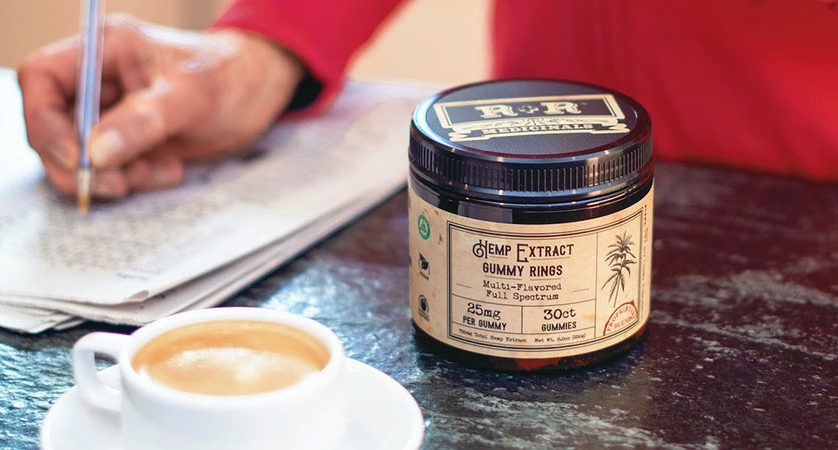 close up view of a person's hand writing on a piece of paper beside a cup of coffee and hemp extract on top of a table